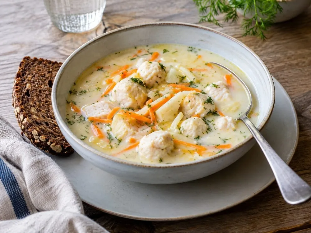 A bowl of Bergen fish soup, pale and creamy, with finely shredded carrot and root vegetables, small fish balls and a spoon resting against the rim.