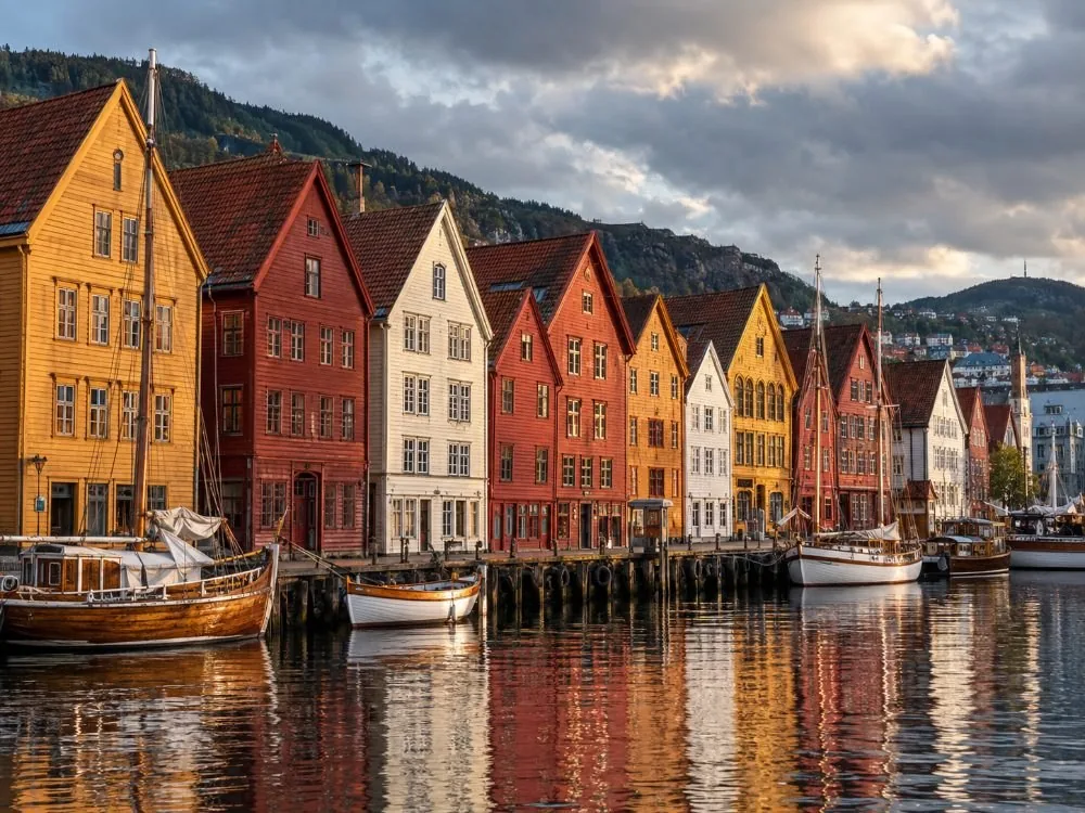 The wooden gable fronts of Bryggen along the harbour in low afternoon light, ochre and red and cream, leaning slightly, with boats tied up in front of them.
